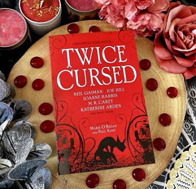 image of a copy of the book Twice Cursed, edited by Marie O'Regan and Paul Kane, lying on a cream cloth surrounded by red beads, pink roses and candles and silver leaves